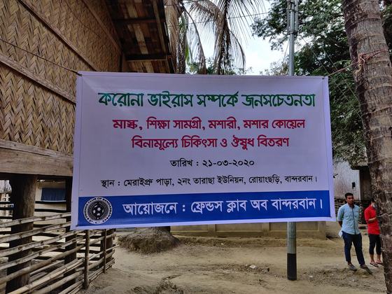 A printed Bengali banner announcing the free medical camp, strung between a bamboo house and a post