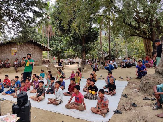 Village children sitting on a tarpaulin listening to the session, with adults seated behind them