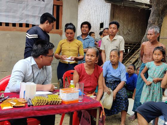 A doctor speaking with a seated patient while villagers stand around the table