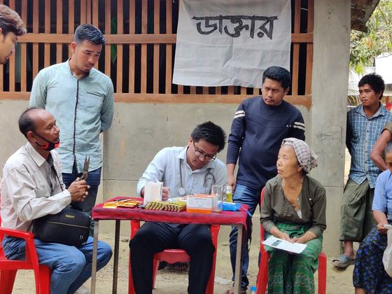 A doctor writing at a red table beneath a hand-lettered cloth sign reading &ldquo;Doctor&rdquo; in Bengali