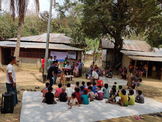Wide view of the camp — a village clearing with tin-roofed houses, a consultation table and people gathered around it