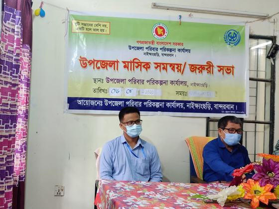 Two masked officials seated beneath a Bengali banner announcing the monthly upazila coordination meeting