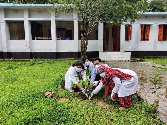 Four health workers in white coats and masks planting a sapling on a grass lawn